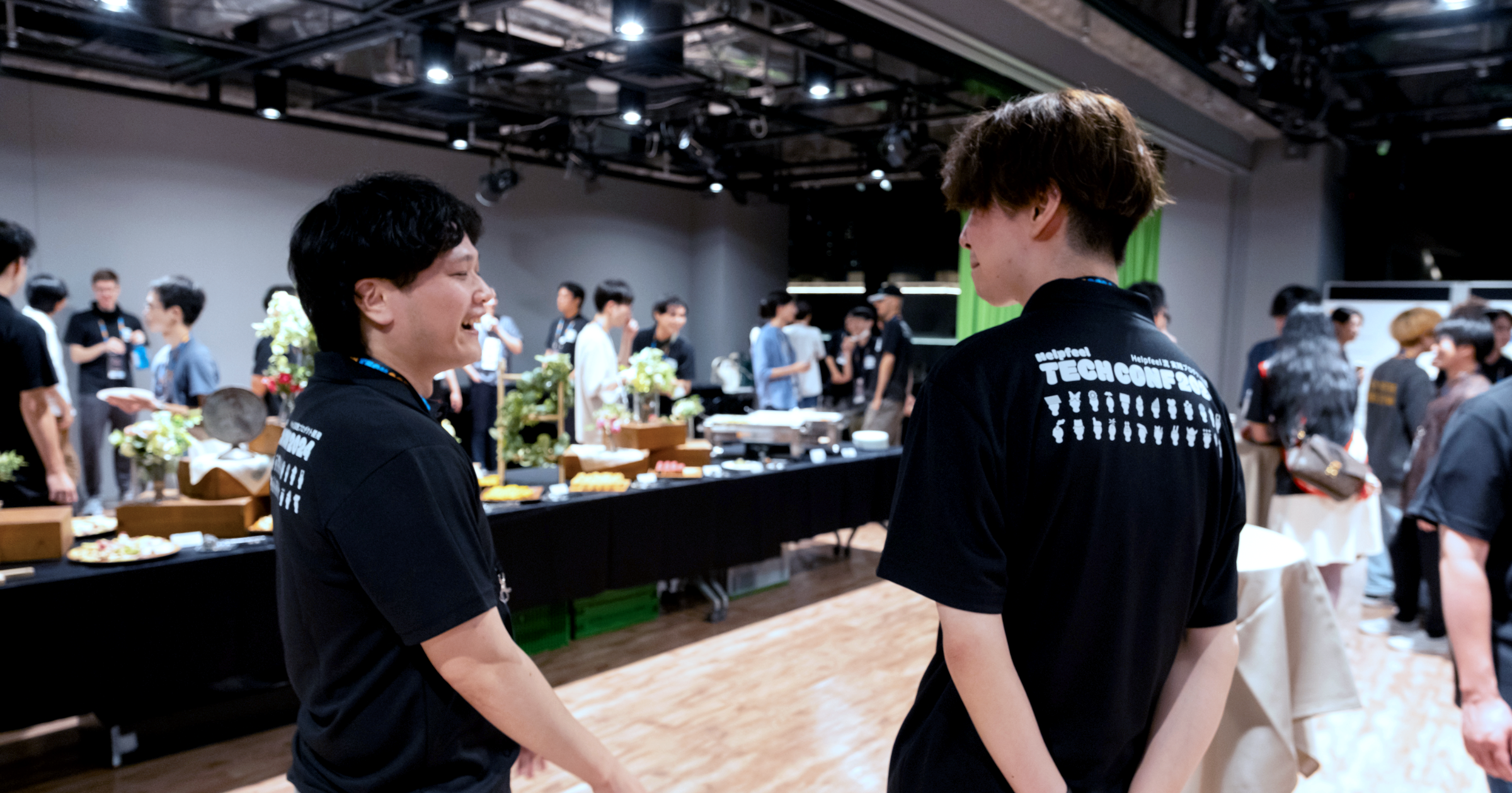 男性二人が向き合って談笑している。奥には長机の上に料理が並んでいる。
